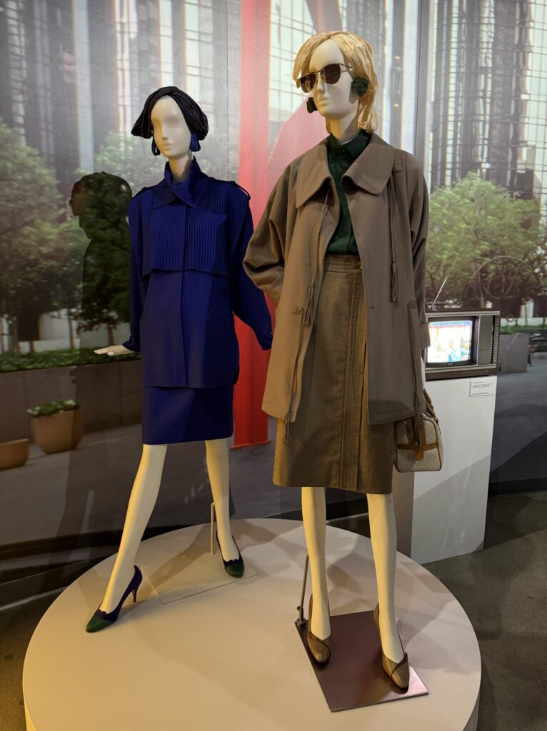 Two mannequins styled in high-powered 1980s workwear: the first wears a gray wool jacket and skirt ensemble by Gucci from Fall/Winter 1984, accessorized with a leather handbag and oversized sunglasses; the second features a bold cobalt blue wool skirt suit with pleated detailing by Bernard Perris from Spring 1986, paired with matching heels and earrings. This fashion is on display courtesy of FIDM at the Totally Awesome Exhibit at the Petersen Automotive Museum. Photograph by Regina Luz Jordan for Hollywoodland News