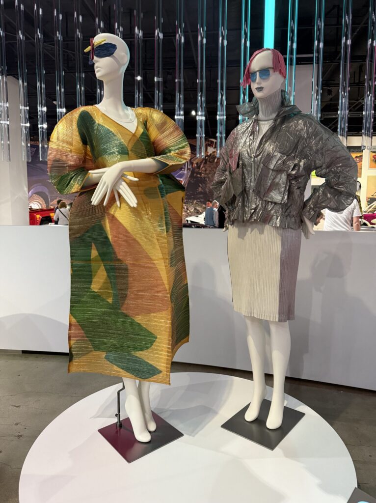 Two mannequins display avant-garde fashion designs by Issey Miyake from the 1990s. The left figure wears the vibrant, sculptural “Pau” coat made of heat-set cellophane in bold orange, green, and yellow geometric patterns, featuring dramatic origami-style sleeves and pleated structure. The right mannequin wears a silver heat-treated polyester jacket with a futuristic sheen and voluminous shape over a simple pleated skirt, reflecting Miyake’s experimental, tech-forward approach to fashion. This fashion is on display courtesy of FIDM at the Totally Awesome Exhibit at the Petersen Automotive Museum. Photograph by Regina Luz Jordan for Hollywoodland News