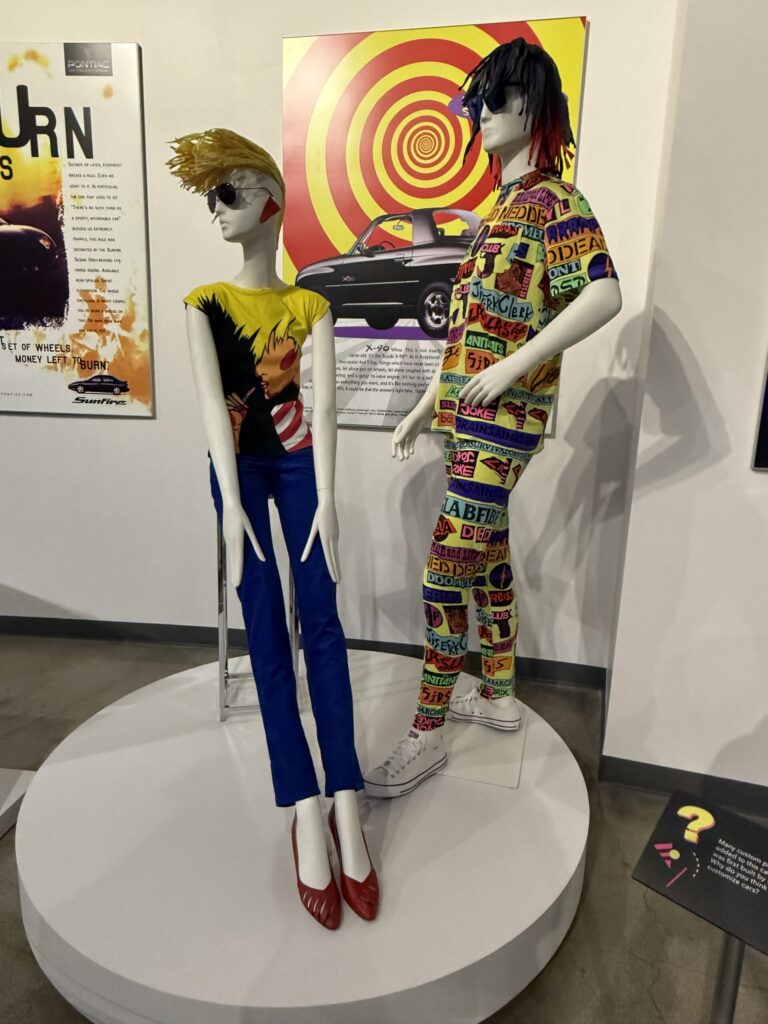 Two mannequins styled in vibrant, punk-inspired 1980s and 1990s fashion. The mannequin on the left wears a black and yellow abstract graphic T-shirt from Theodore Boutique (1980) paired with electric blue Fiorucci corduroy pants featuring Memphis-style designs, and red heels. The mannequin on the right sports a full printed ensemble from Stephen Sprouse’s Spring/Summer 1998 collection, covered in colorful, stenciled neon graffiti-style text. Both outfits exemplify the bold, rebellious fusion of high fashion and streetwear from the post-punk and new wave scenes. This fashion is on display courtesy of FIDM at the Totally Awesome Exhibit at the Petersen Automotive Museum. Photograph by Regina Luz Jordan for Hollywoodland News