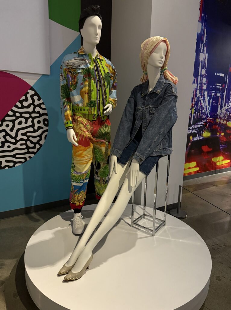 A mannequin duo styled in quintessential ’90s flair. The left mannequin wears a vivid Gianni Versace Spring/Summer 1993 cotton jacket and pants set featuring maximalist Miami-inspired tropical and aquatic prints in neon tones. The right mannequin sports a double denim look with a rare 1994 Yves Saint Laurent for Levi Strauss jacket paired with a 1985–90 Guess? by Georges Marciano denim skirt, embodying the rebellious, high-fashion spirit of ‘90s youth culture. The scene is set against a backdrop of pop art shapes and neon city lights, echoing the boldness of the era. This fashion is on display courtesy of FIDM at the Totally Awesome Exhibit at the Petersen Automotive Museum. Photograph by Regina Luz Jordan for Hollywoodland News