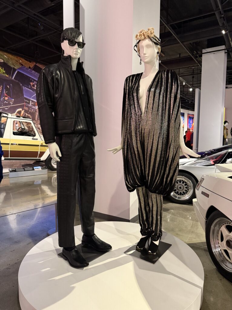 from 1983, styled with sunglasses and sleek hair; the other in a shimmering black-and-silver striped jumpsuit by Norma Kamali, circa 1982, featuring dramatic draping and a plunging neckline, topped with a floral headpiece. This fashion is on display courtesy of FIDM at the Totally Awesome Exhibit at the Petersen Automotive Museum. Photograph by Regina Luz Jordan for Hollywoodland News